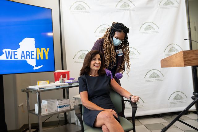 Gov. Kathy Hochul, smiling sitting in a chair, as she's being administered a COVID-19 booster shot